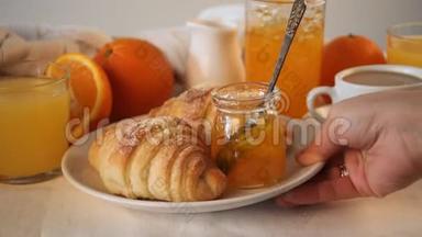女人的手在桌子上放了一个盘子，上面放着新鲜的羊角面包。 附近有橘子，橘子果酱和果汁。 早餐概念。